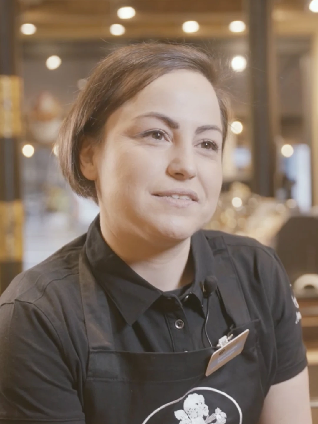 Femme portant un uniforme noir avec un micro et un badge, dans un environnement intérieur lumineux.
