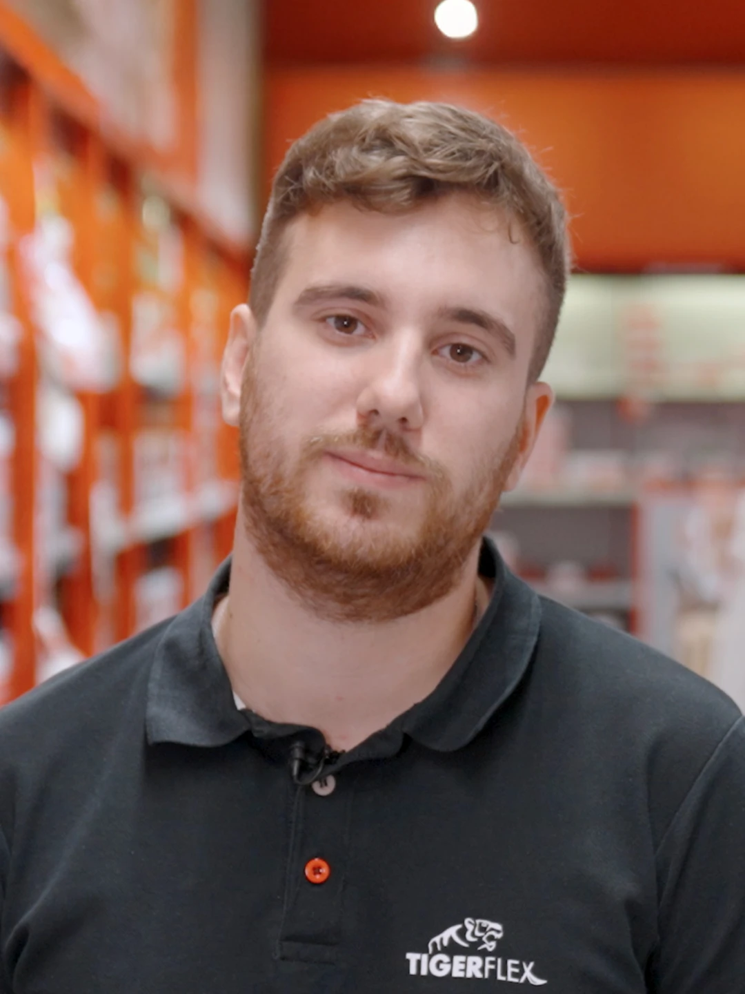 Jeune homme avec barbe et cheveux courts portant un polo noir avec logo Tigerflex, dans un magasin aux étagères rouges floues en arrière-plan.