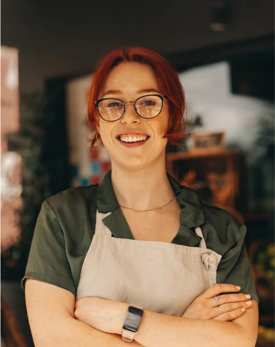 Jeune femme rousse souriante portant des lunettes, un tablier beige et une montre connectée, bras croisés dans un intérieur flou.