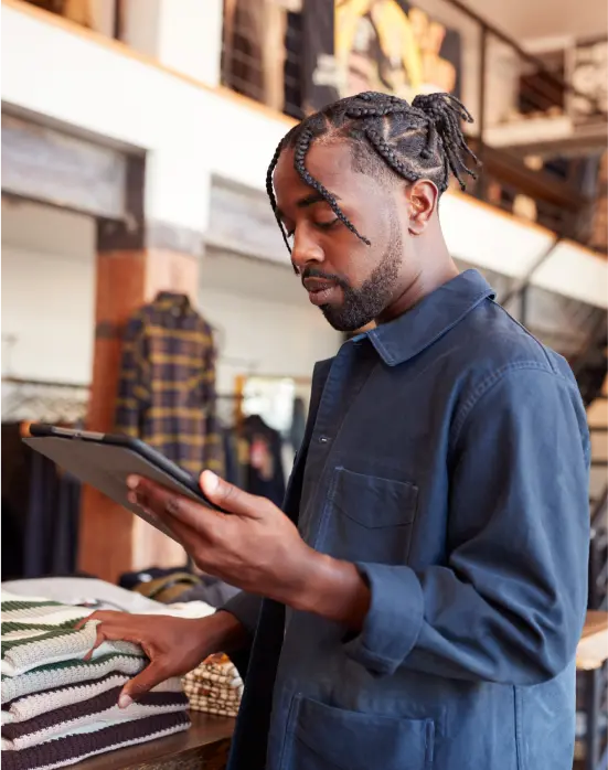 Homme avec dreadlocks et veste bleue utilisant une tablette tout en touchant un empilement de pulls pliés dans un magasin.