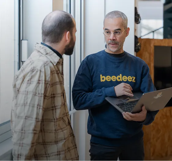 Deux hommes discutent, l'un tenant un ordinateur portable et portant un sweat-shirt bleu avec le logo 'beedeez'.