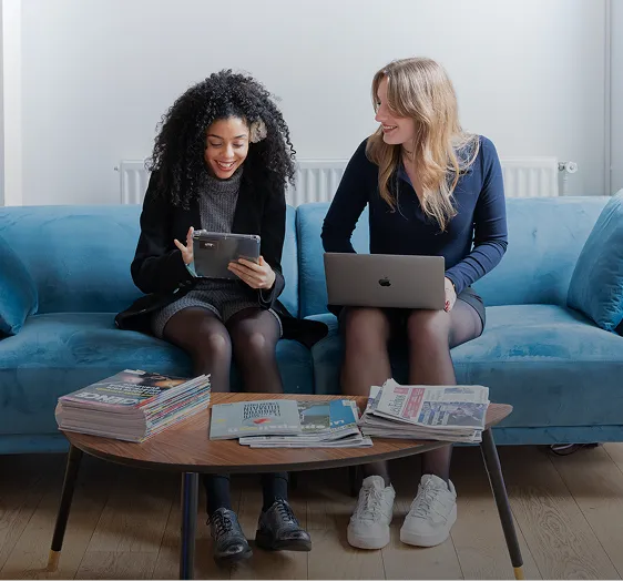 Deux femmes assises sur un canapé bleu, l’une utilisant une tablette et l’autre un ordinateur portable, devant une table basse avec des magazines et journaux.